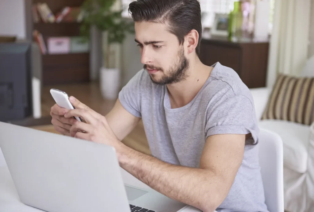 Uomo seduto davanti al portatile che cerca qualcosa sullo smartphone