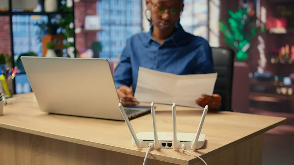 Donna davanti al portatile con un foglio in mano, router Wi-Fi in primo piano sulla scrivania