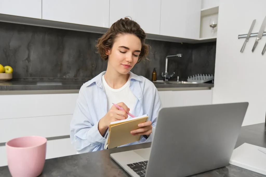 Donna al tavolo di cucina con il portatile aperto che prende appunti su un quaderno